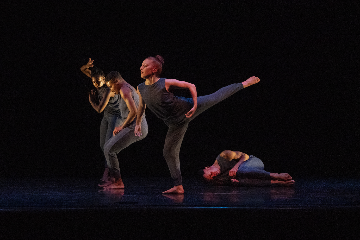 The picture is four dancers in a tableau. From left to right: two dancers look down, their shoulders hunched; one dancer extends her leg in arabesque; another dancer is prone on the floor in the fetal position.